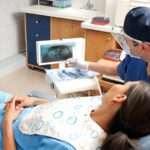 Woman in dental chair with man in background.